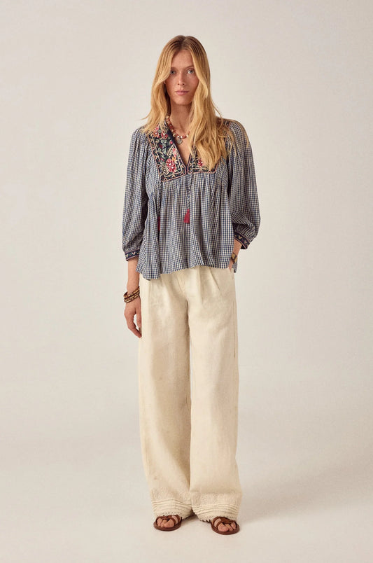 Model wearing the April Blouse in Bleu Picnic, a blue and white gingham cotton blouse with floral embroidered yoke, tassel ties, long sleeves, and babydoll silhouette paired with ivory wide-leg pants.