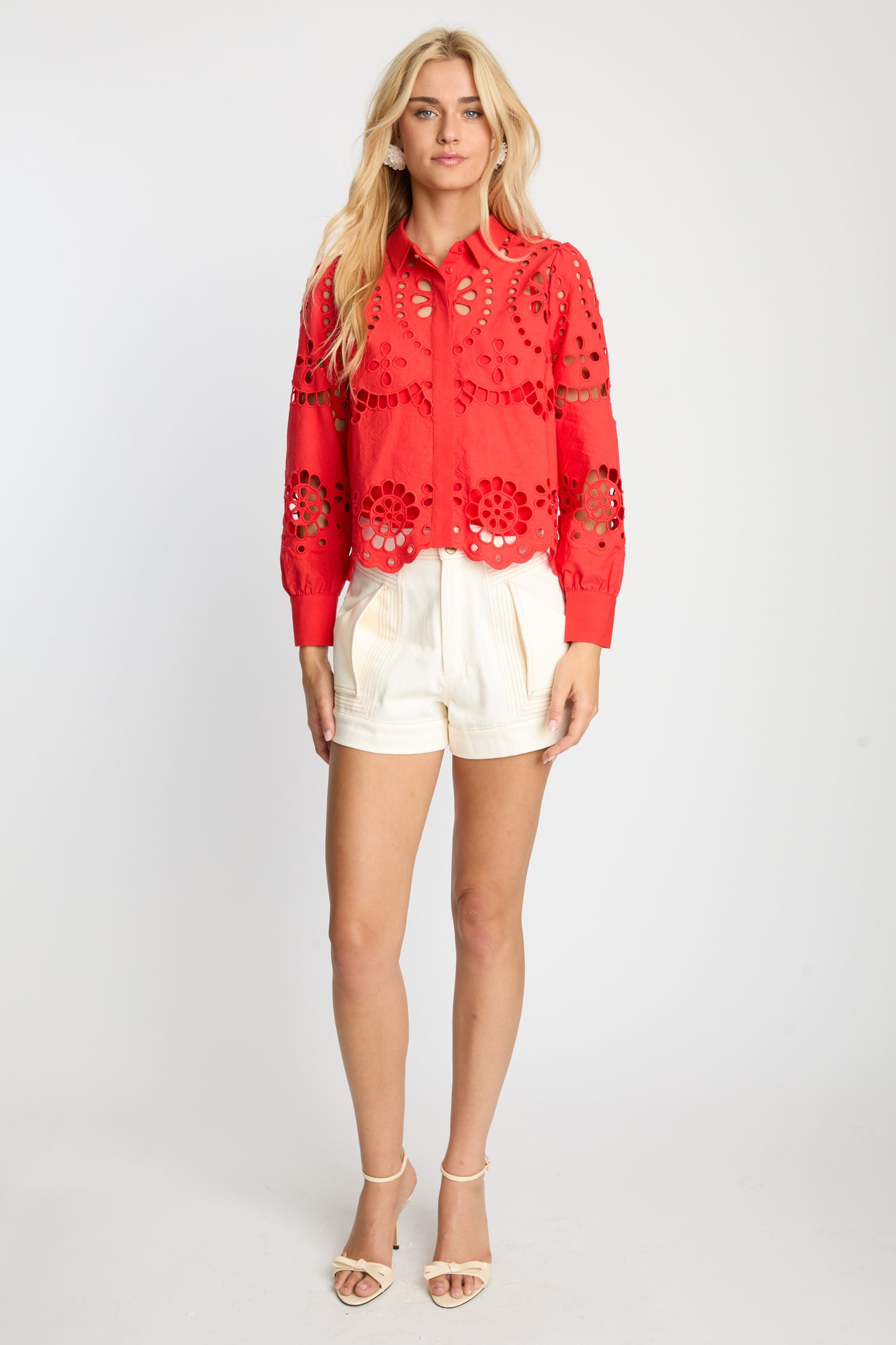 Model wearing the Emmie Top in Poppy Red Eyelet Lace, a cropped red cotton blouse with intricate eyelet embroidery, scalloped hem, button front, and puffed long sleeves paired with cream shorts.