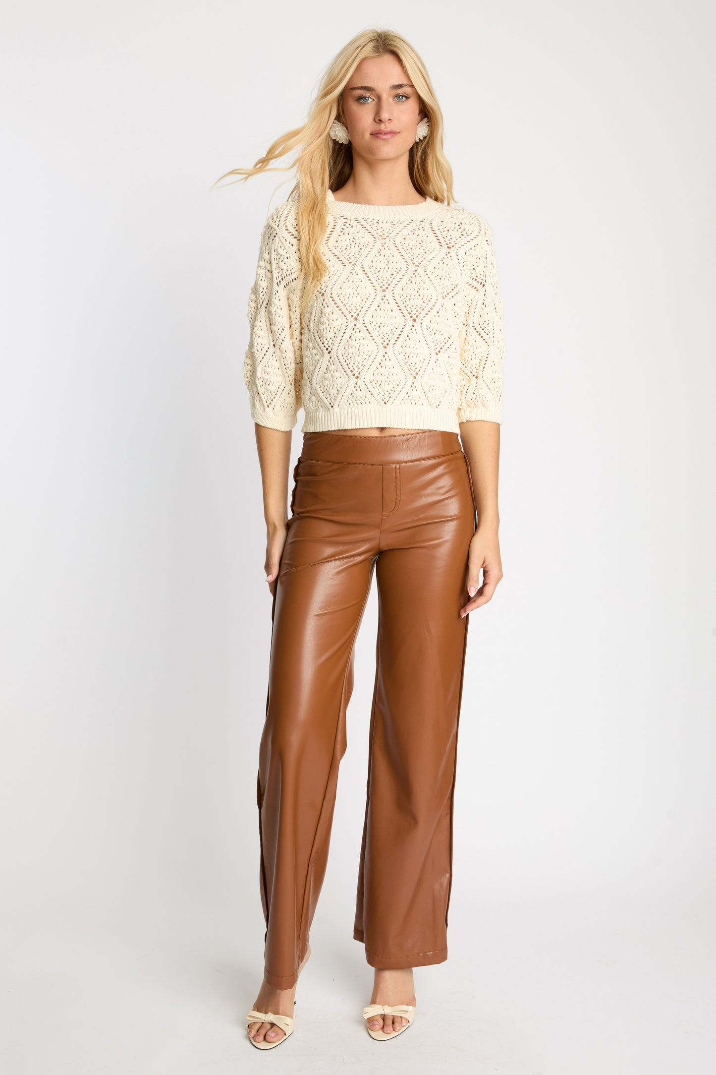 Model wearing the Layla Sweater in cream featuring a cropped openwork knit design with diamond patterns, three-quarter puff sleeves, and a round neckline paired with camel faux leather pants.