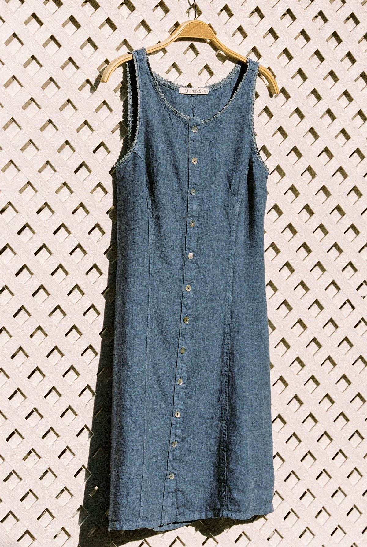 The Linen Pinafore in Blueberry displayed hanging against a white lattice backdrop.