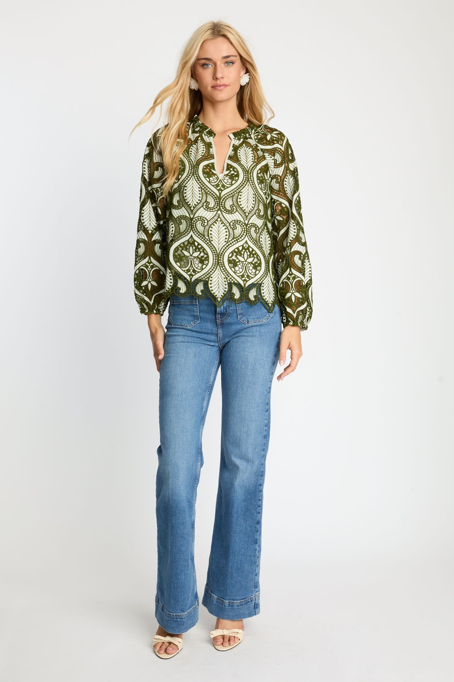 Model wearing the Miki Top in loden green featuring intricate embroidered eyelet fabric, paisley-inspired cut-out detailing, split V-neckline, long sleeves, and scalloped hem paired with blue jeans.
