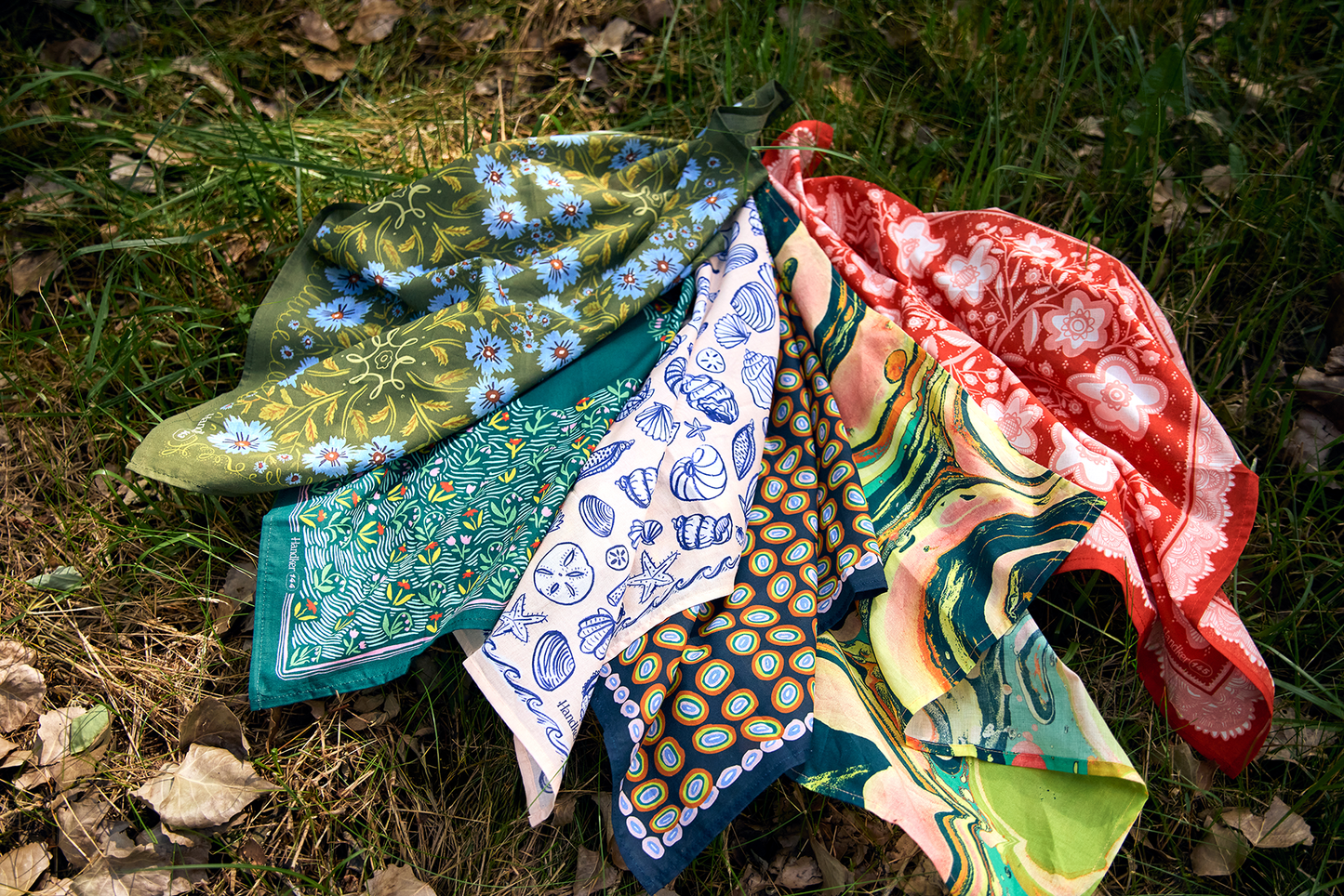 Assorted colorful floral bandanas, including the red-orange Lacey Bandana, arranged outdoors on the grass.