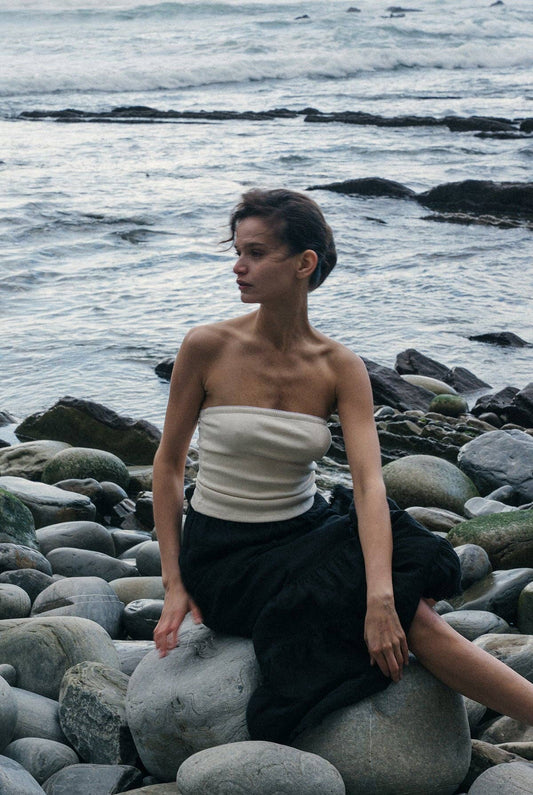 Model wearing the Pointelle Tube Top in Creme with a black flowing skirt while seated on coastal rocks by the ocean.