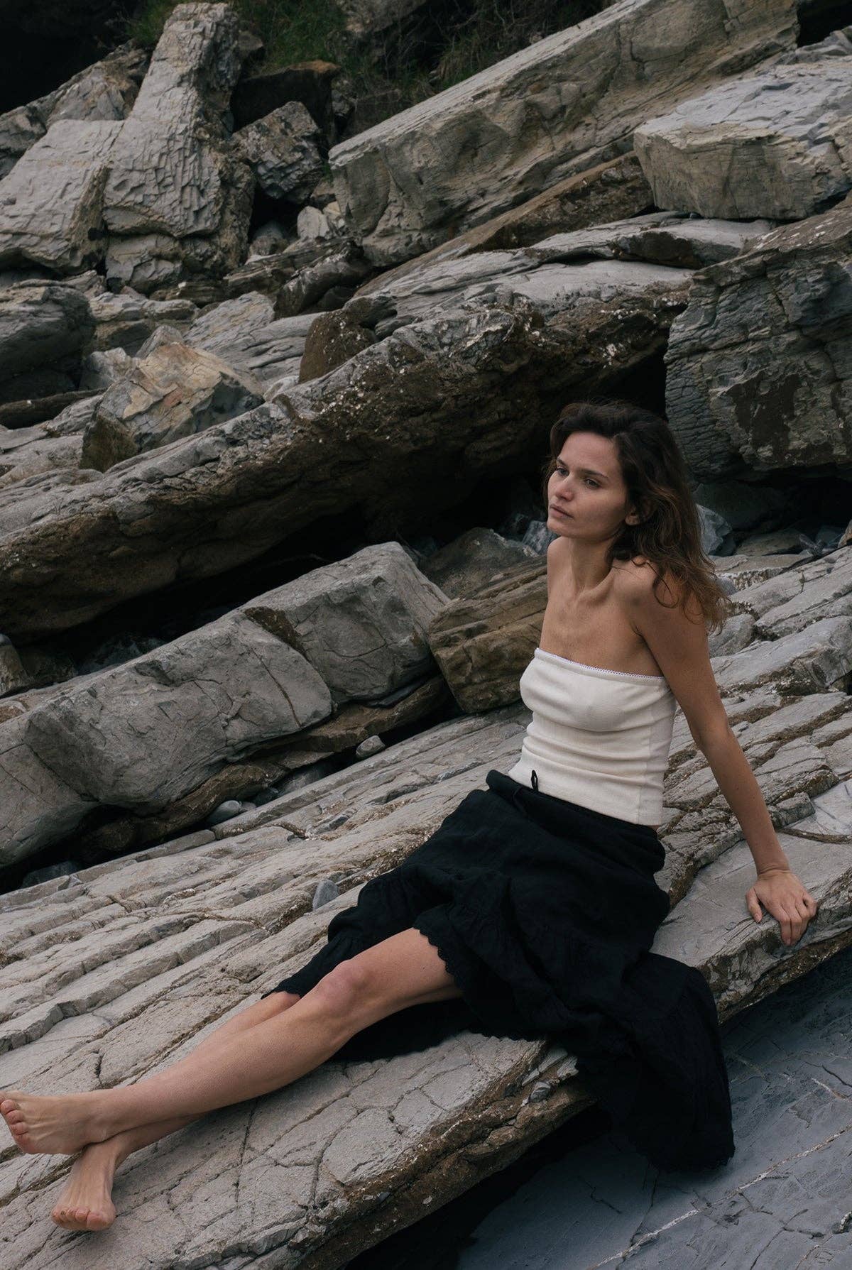 Model wearing the creme pointelle tube top styled with a black skirt while seated against layered coastal rocks.