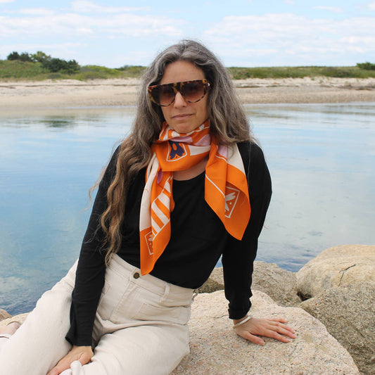 Adult model sitting by the beach wearing the Soar & Sing silk scarf tied around the neck with a black top and neutral pants.