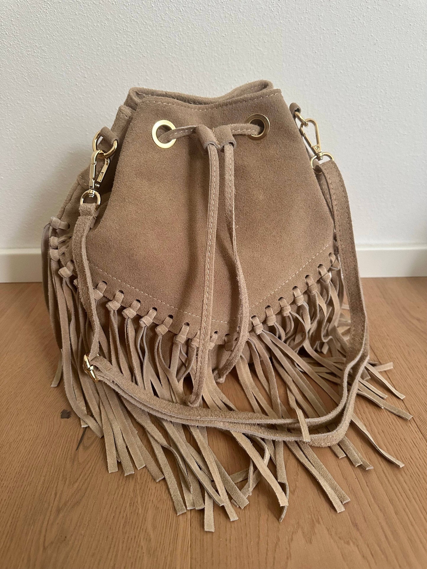 Taupe suede bucket bag with long fringe detailing, drawstring closure, adjustable shoulder strap, and gold-tone hardware, handcrafted in Italy.
