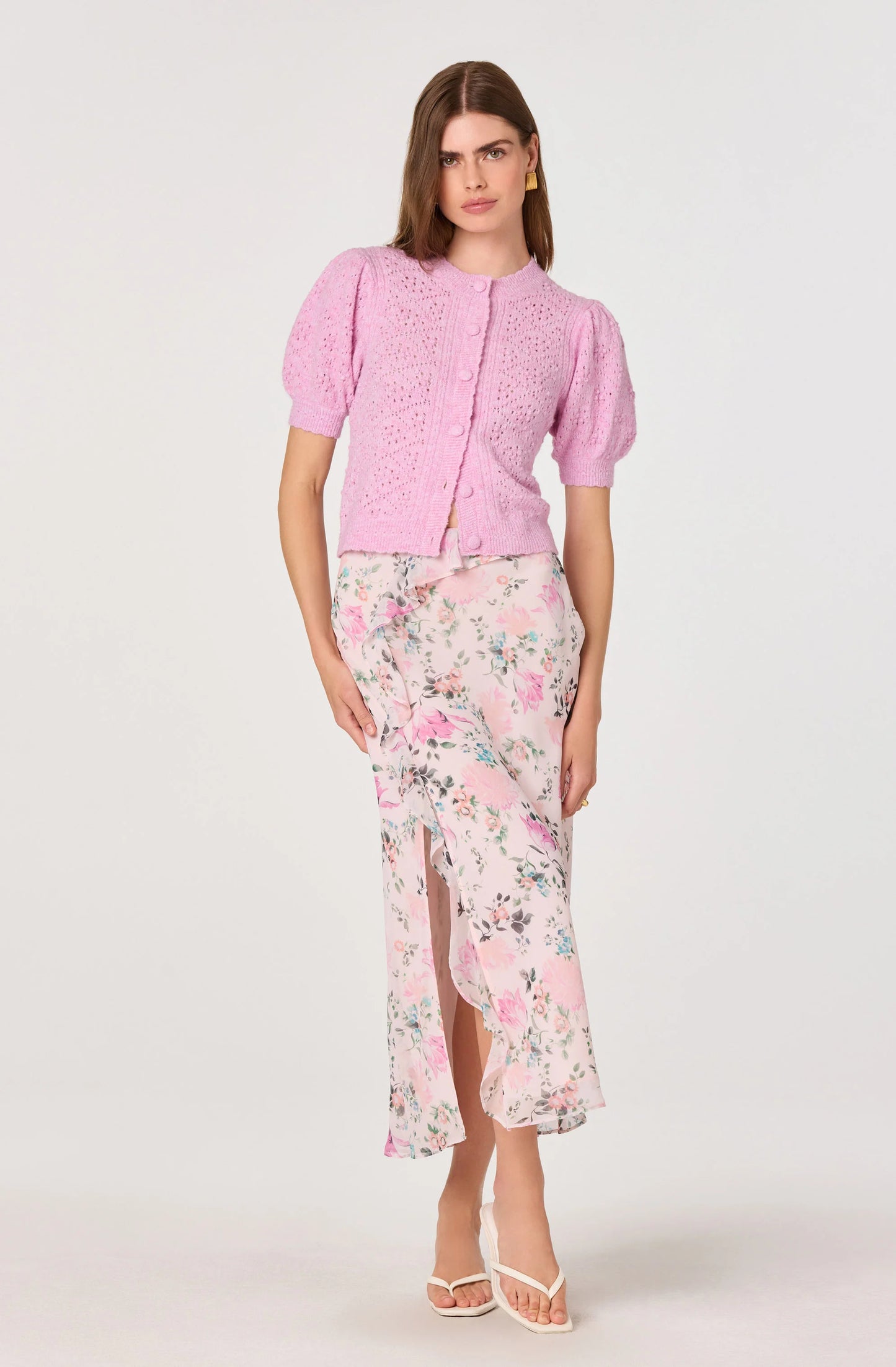 Model wearing the Ananda Floral Bias-Cut Ruffle Midi Skirt in a blush floral print with a ruffle detail and front side slit, styled with a pink cardigan.
