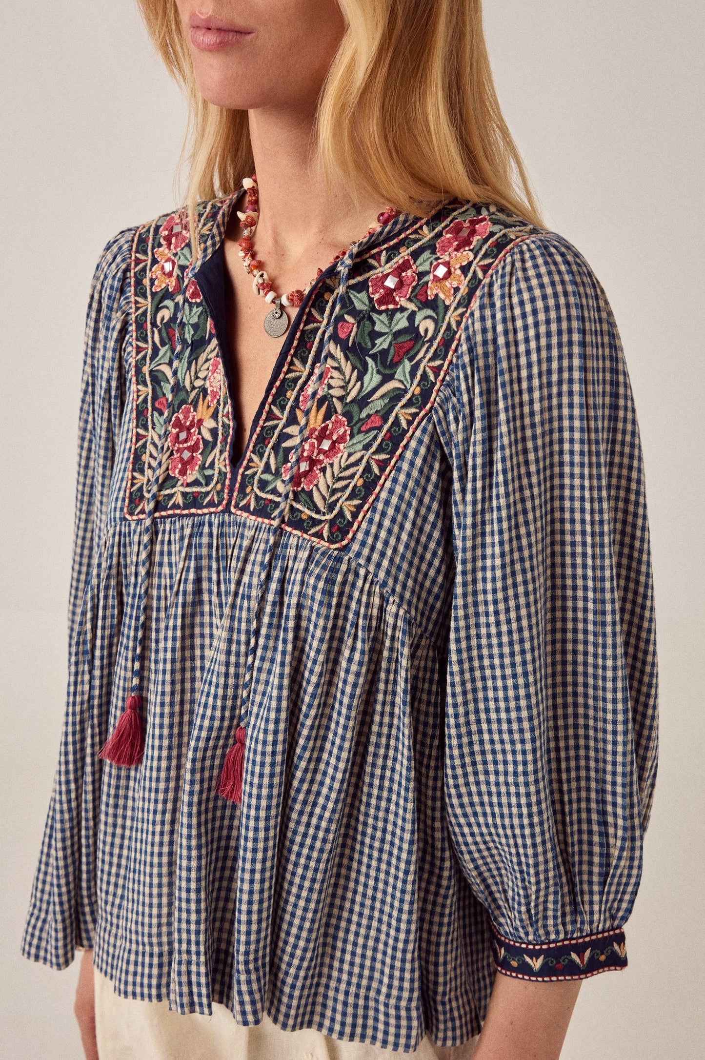 Close-up of the April Blouse in Bleu Picnic highlighting intricate floral embroidery, blue and white gingham cotton fabric, gathered bodice, and tassel tie neckline.
