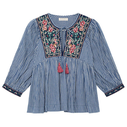 Flat lay front view of the April Blouse in Bleu Picnic featuring blue and white gingham cotton fabric, floral embroidered yoke, split V-neck with pink tassel ties, and embroidered cuffs.