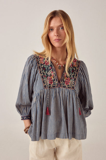 Front view of the April Blouse in Bleu Picnic featuring blue gingham check crepe fabric, floral embroidery on the yoke, split V-neckline, and pink tassel tie details.