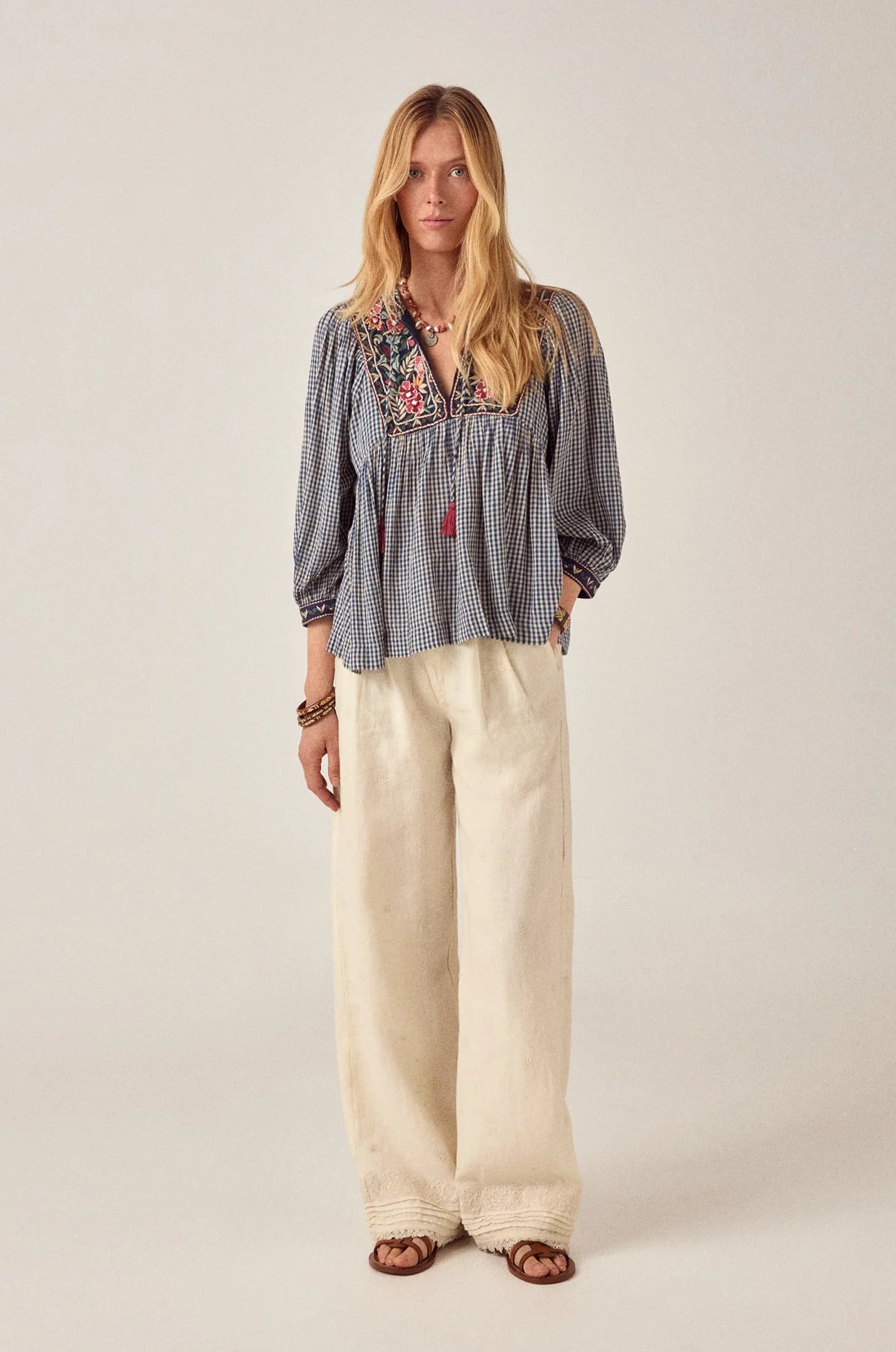 Model wearing the April Blouse in Bleu Picnic, a blue and white gingham cotton blouse with floral embroidered yoke, tassel ties, long sleeves, and babydoll silhouette paired with ivory wide-leg pants.