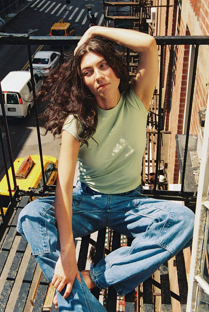 Model seated outdoors wearing the Pistachio Bebe Organic Tee with relaxed blue jeans.