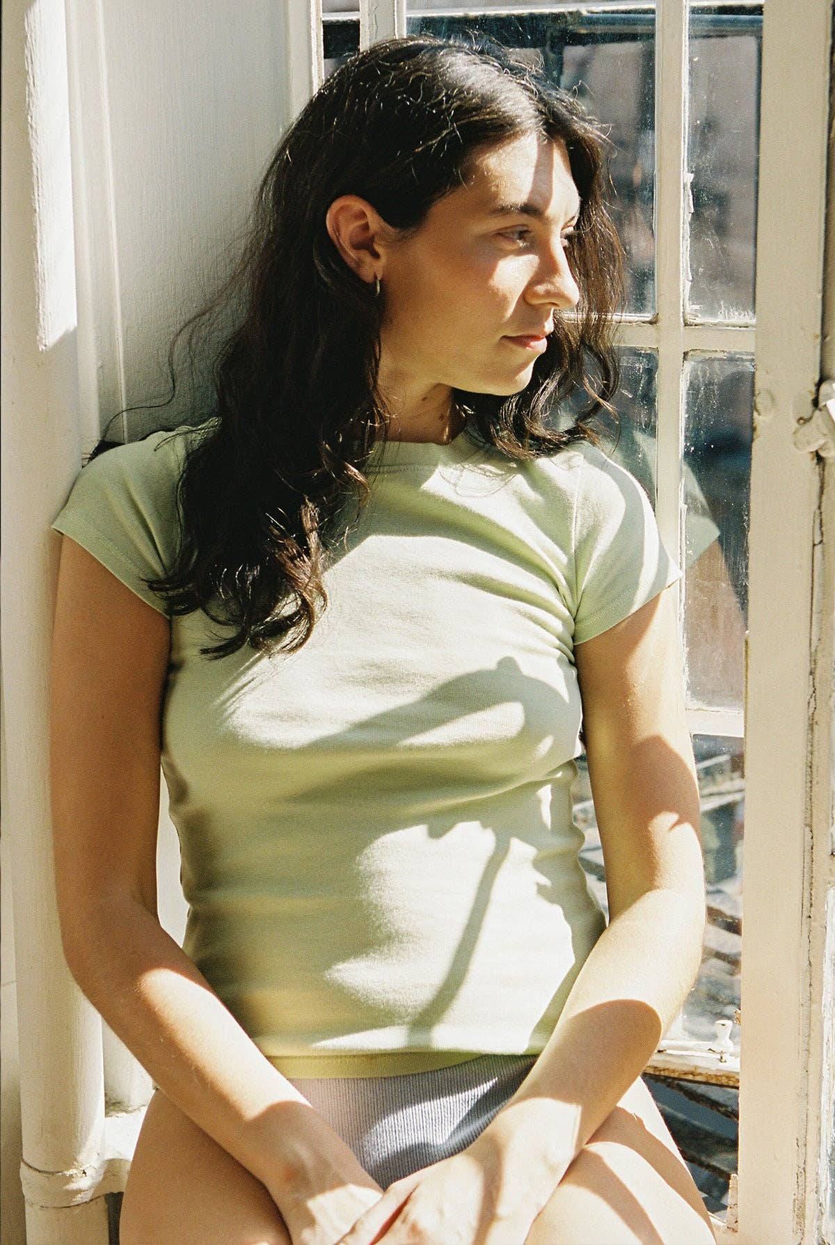 Seated model wearing the fitted Pistachio Bebe Organic Tee beside a sunlit window.