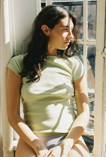 Seated model wearing the fitted Pistachio Bebe Organic Tee beside a sunlit window.