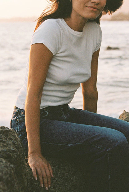 Model seated by the water wearing the Bebe Organic Teen Tee in White with dark wash denim.