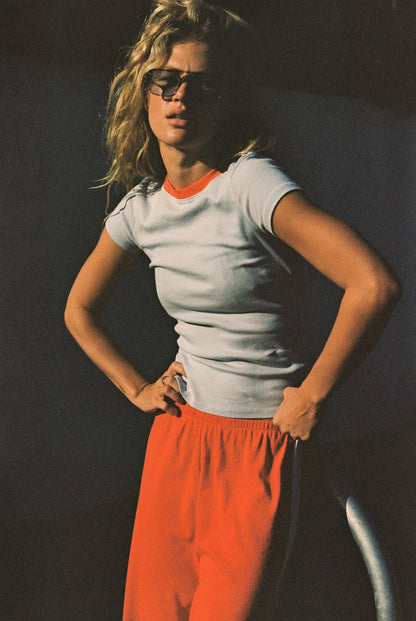 Model wearing the light gray Bebe Ringer Tee with coral neckline trim and orange trousers.