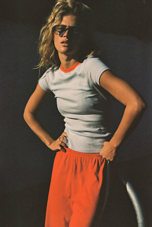 Model wearing the light gray Bebe Ringer Tee with coral neckline trim and orange trousers.