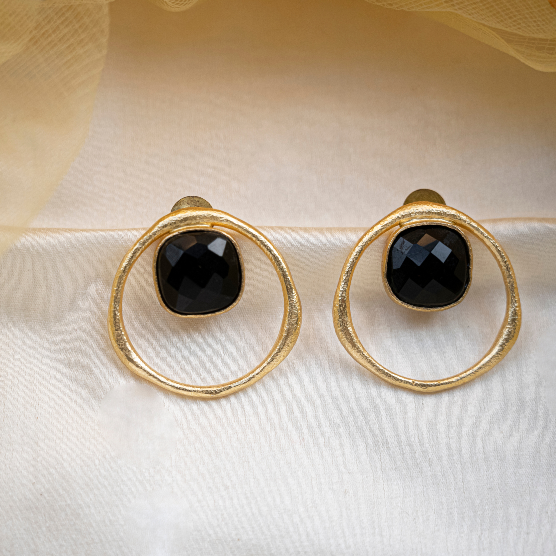 Pair of handmade black onyx stud earrings with sculptural 18k gold-plated circular frames displayed on ivory fabric.