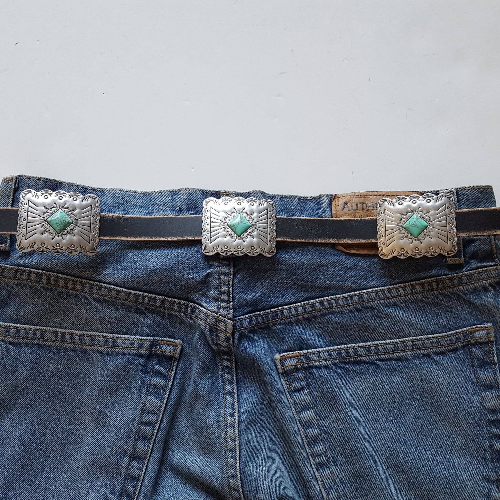Black genuine distressed leather belt featuring engraved rectangular conchos with turquoise stone details styled on denim jeans