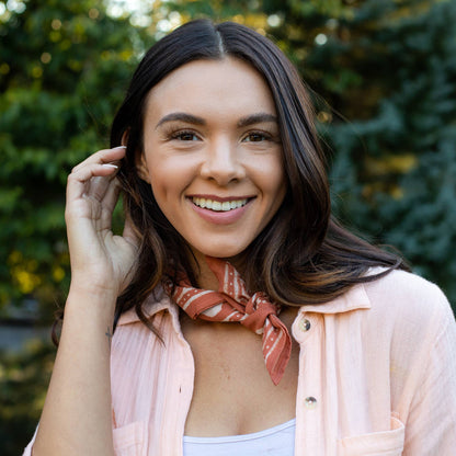 Woman wearing a burnt orange block printed cotton bandana tied around her neck. The handcrafted bandana features a tile grid print and geometric border, styled with a light pink button-up shirt outdoors.