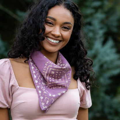 Woman wearing a mauve pink block printed cotton bandana tied around her neck. The handcrafted bandana features a geometric diamond motif with an ivory decorative border, styled with a soft pink top outdoors.