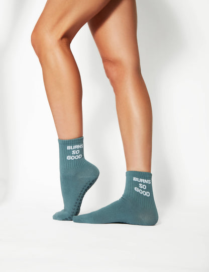 Close-up of blue BURNS SO GOOD grip socks showing ribbed cuffs, slogan detail, and non-slip grip dots on the sole.