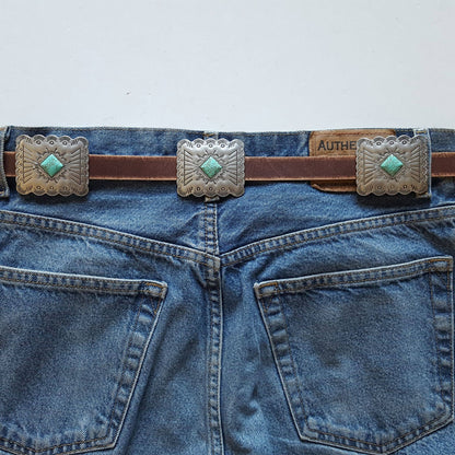 Brown genuine distressed leather belt with engraved rectangular silver conchos and turquoise stone accents styled on denim jeans back view