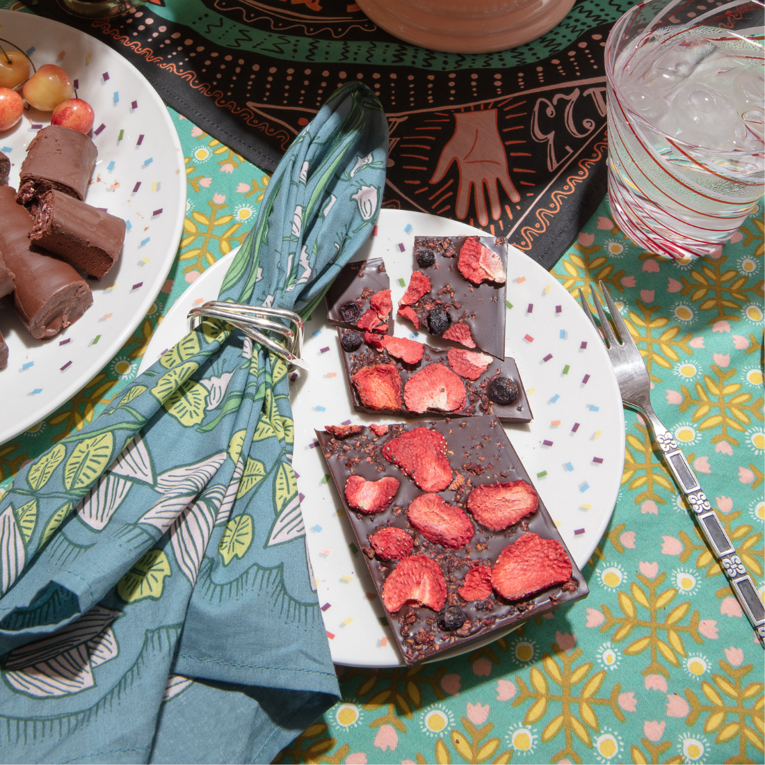 Teal floral Cassie bandana styled on a table with dessert plates, chocolate, cherries, and a glass of ice water.