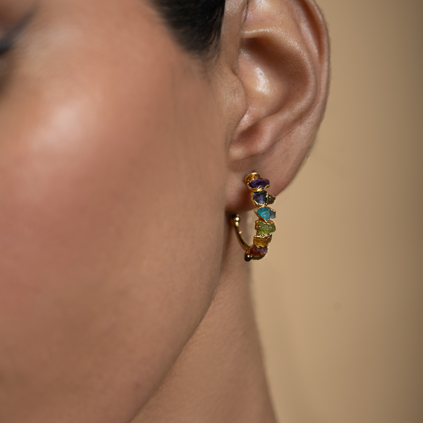 Close-up of a gold-plated chakra hoop earring with multicolored natural stones worn on the ear.