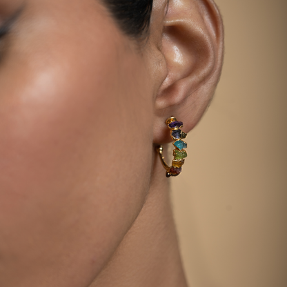 Close-up of a gold-plated chakra hoop earring with multicolored natural stones worn on the ear.