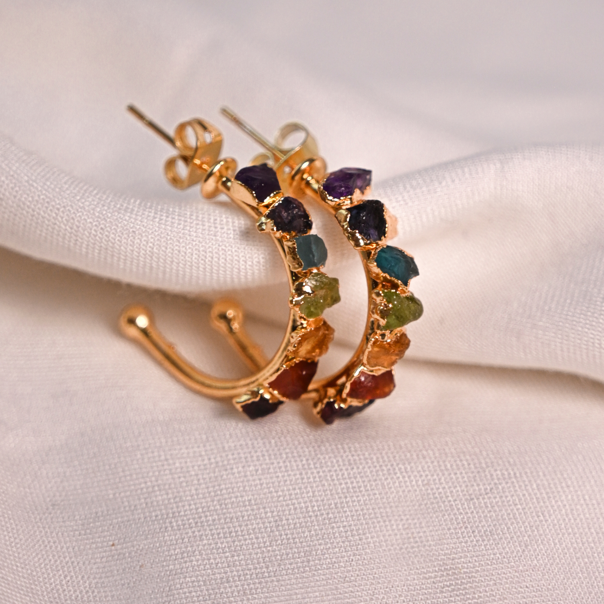 Gold-plated chakra hoop earrings set with colorful raw natural stones on soft white fabric.
