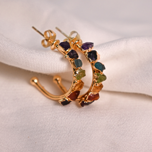Gold-plated chakra hoop earrings set with colorful raw natural stones on soft white fabric.