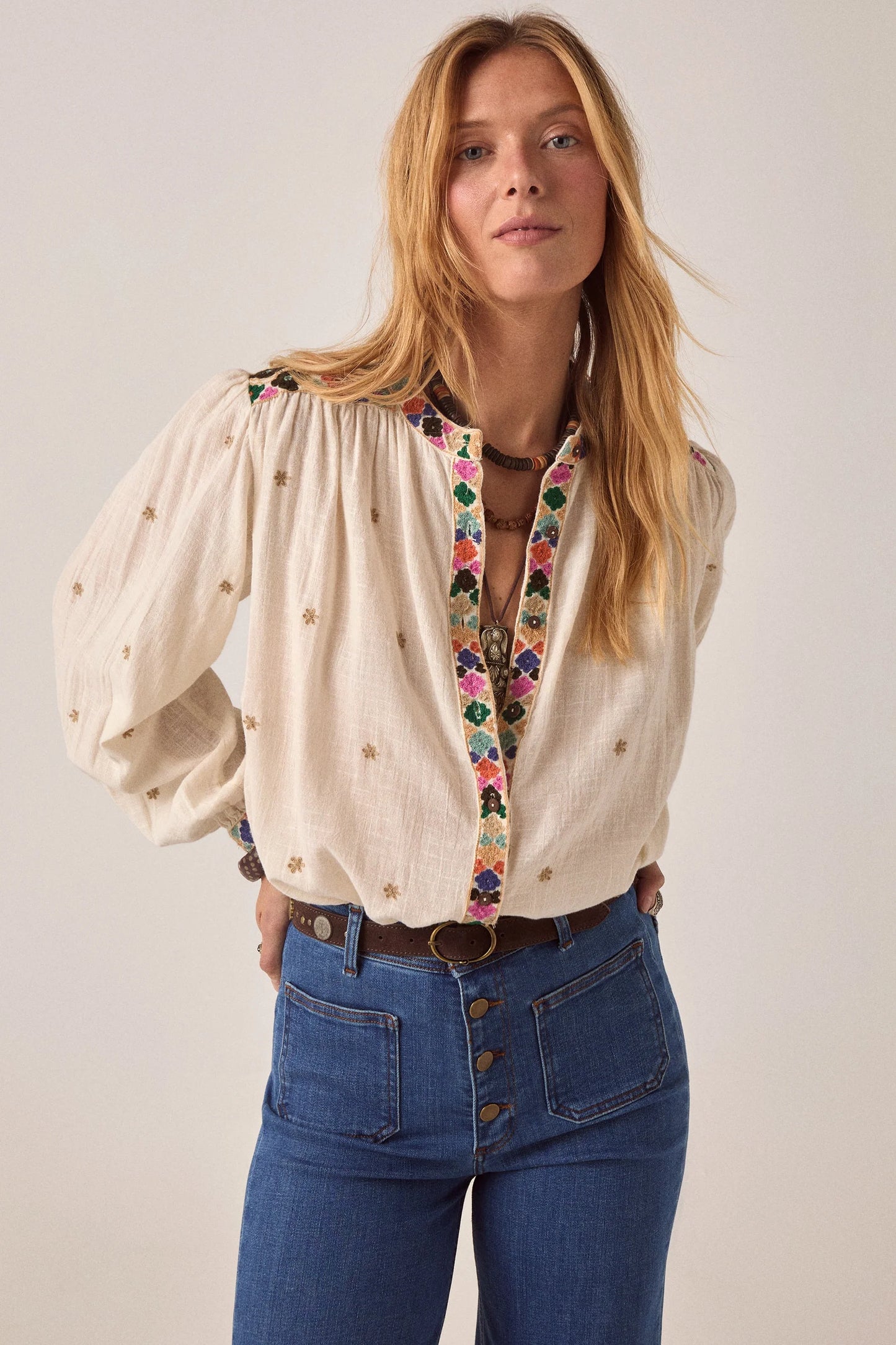 Model wearing the Cleo blouse in off-white with multicolor embroidered trim, gathered neckline, and long balloon sleeves paired with dark denim jeans.