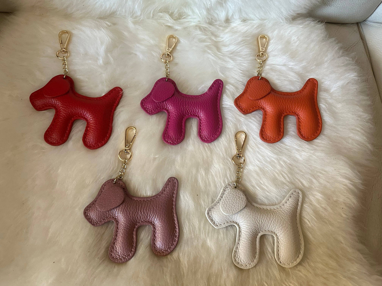 Assorted dog-shaped genuine leather keychains in red, fuchsia, orange, mauve, and cream with gold-tone hardware displayed on a white faux fur surface.
