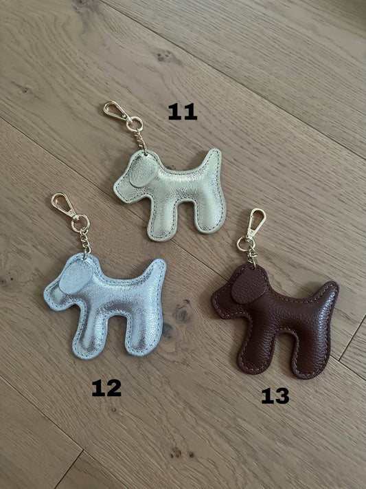 Metallic silver dog-shaped genuine leather keychain with a light gold-tone clasp and chain, handcrafted in Italy and displayed alongside coordinating color options.