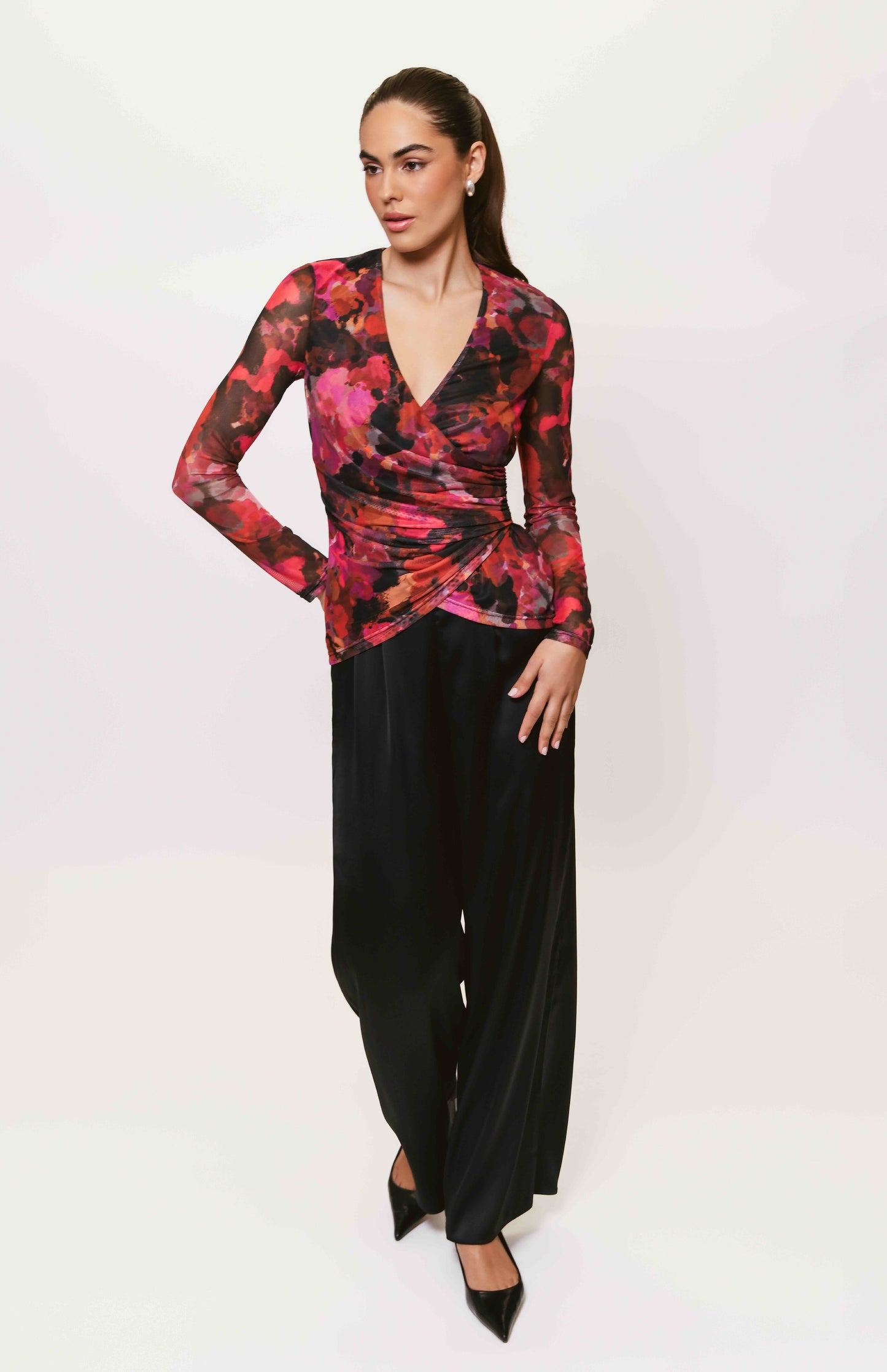 Model wearing the Evia Top in a vibrant red, pink, and black abstract floral print with a ruched faux-wrap design, long sleeves, and V-neckline paired with black satin wide-leg pants.