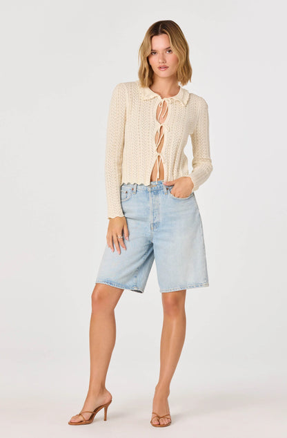 Full-body view of the cream Fannie Cardigan with long sleeves, tie-front detail, and lightweight open-knit fabric styled with denim shorts and heels.