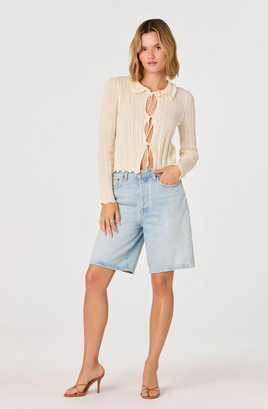 Full-body view of the cream Fannie Cardigan with long sleeves, tie-front detail, and lightweight open-knit fabric styled with denim shorts and heels.