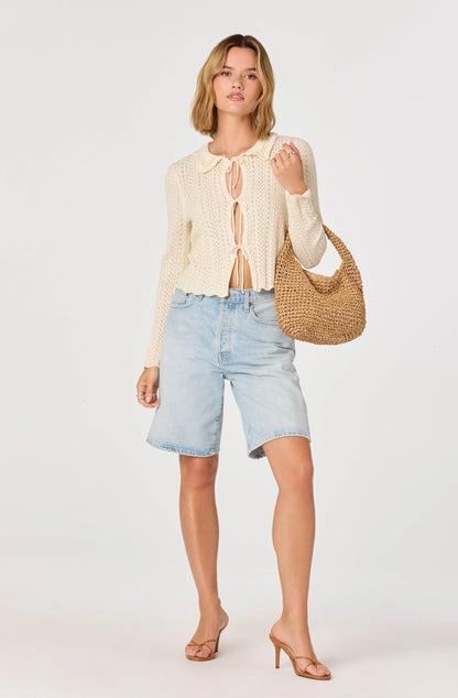 Model wearing the cream Fannie Cardigan with tie-front closure and open-knit texture styled with denim shorts and a woven shoulder bag.