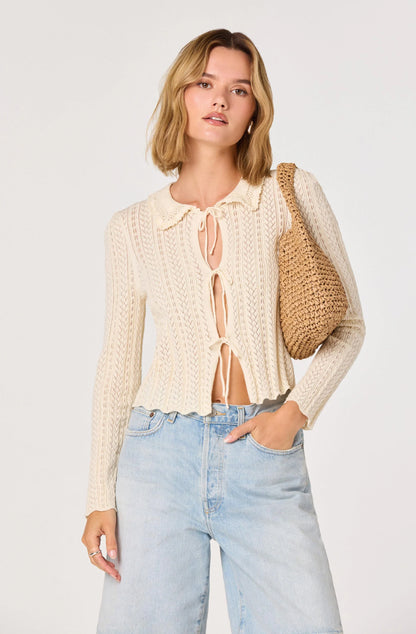 Model wearing the cream Fannie Cardigan featuring an open-knit texture, collared neckline, and tie-front closure paired with light wash denim shorts.