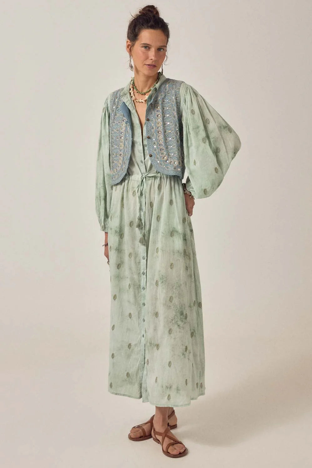 Model wearing the Freya Dress in Mineral Waves in jade cotton voile with tie-dye and paisley print, styled with a blue embroidered vest and sandals.