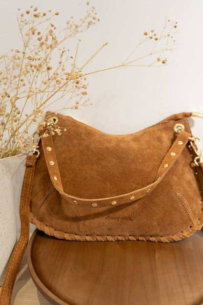 Joëlle camel suede handbag styled on a neutral wooden surface beside delicate dried flowers.