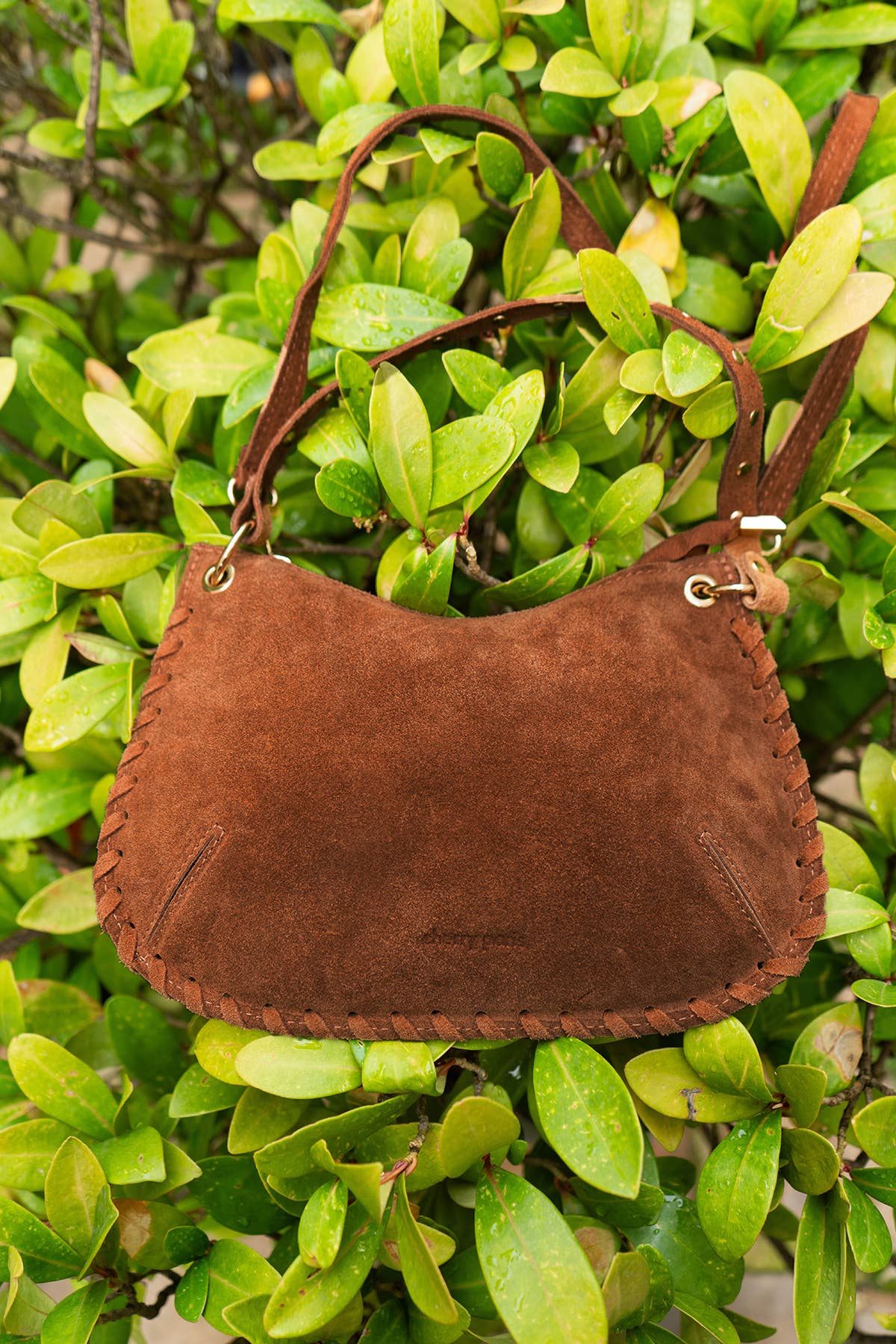 Brown Joelle Suede Bag photographed outdoors against green foliage, showing its soft suede texture and compact shape.