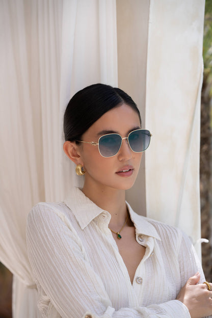 Model wearing Lea women's round sunglasses with a gold-tone frame and gradient gray lenses paired with a white textured blouse and gold accessories.