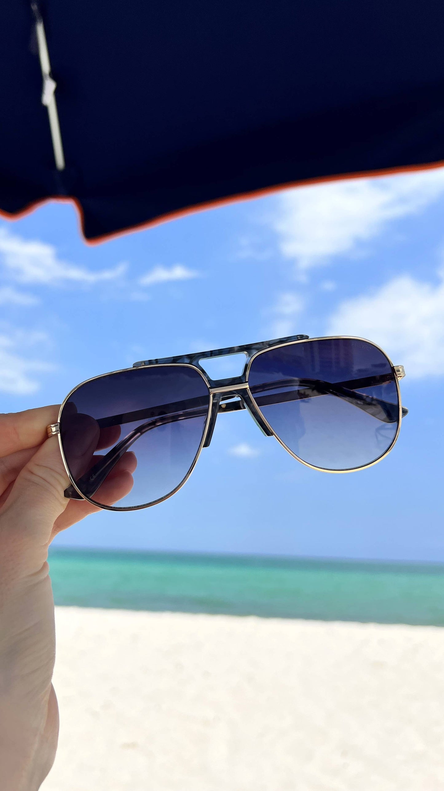 Logan acetate aviator unisex sunglasses shown outdoors at the beach with a light gold frame and gray lenses.