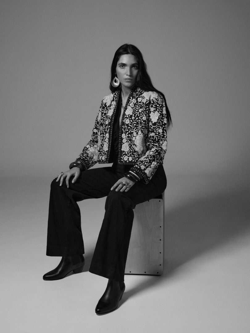 Model wearing the Lola Jacket in Flores floral print with black pants and boots in a seated studio portrait.
