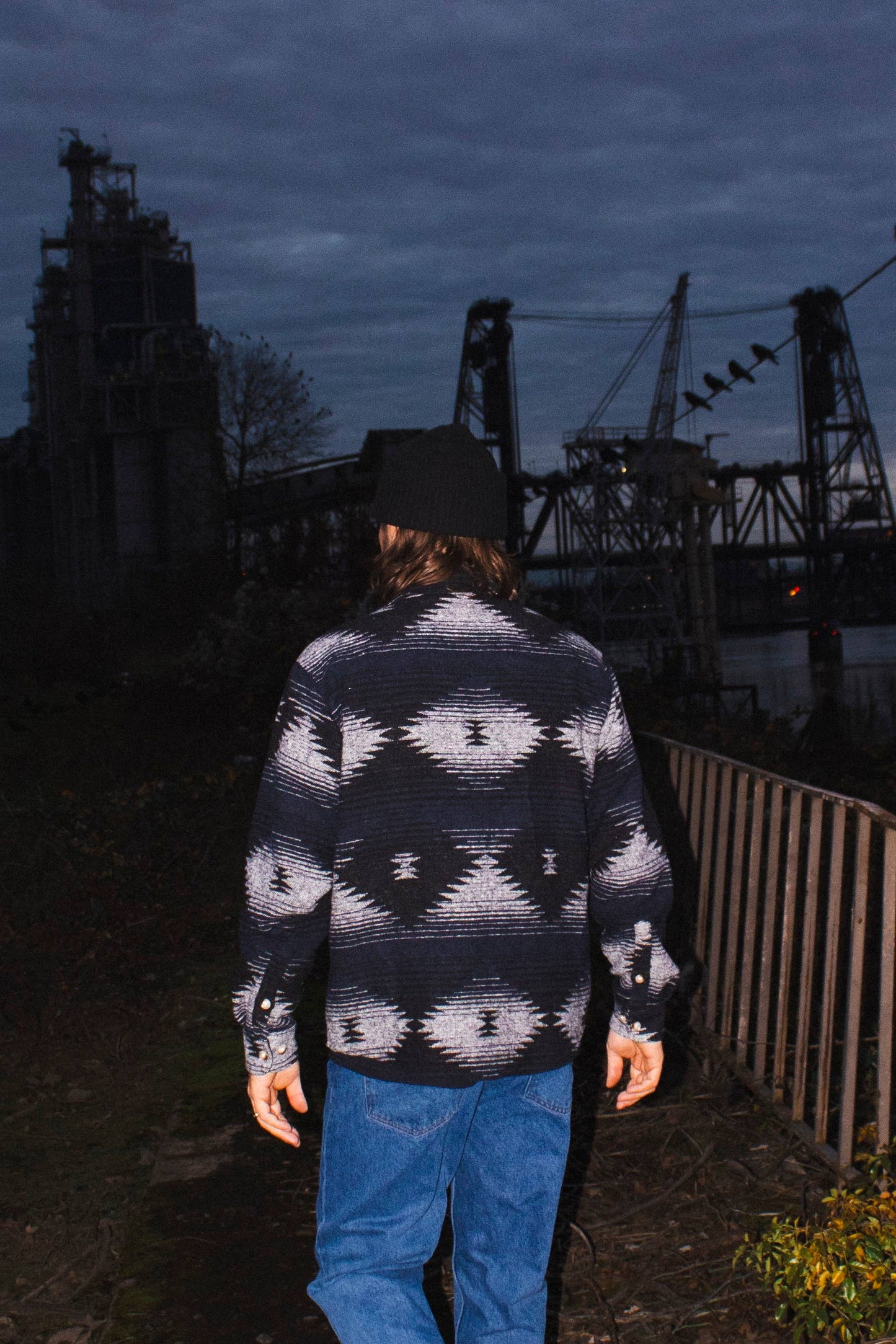 Rear outdoor view of a model wearing the black and blue Moab Mid-Weight Button Down near an industrial riverside setting.