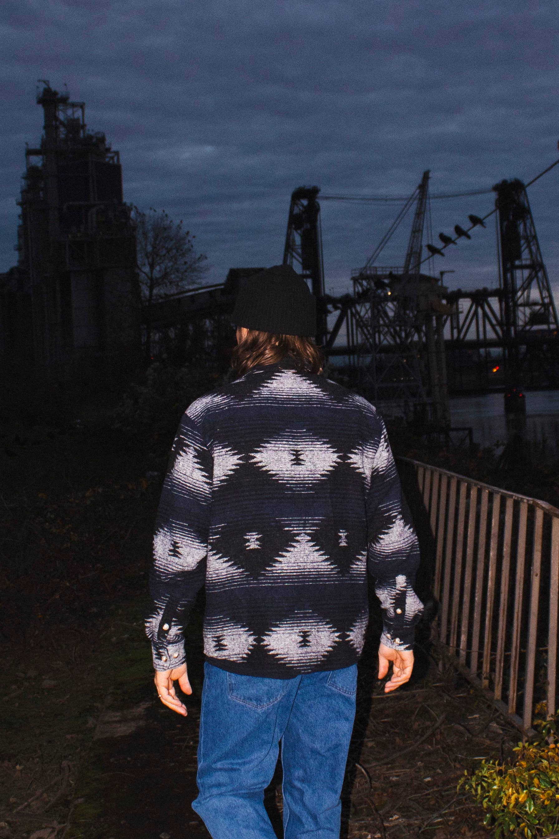 Rear outdoor view of a model wearing the black and blue Moab Mid-Weight Button Down near an industrial riverside setting.