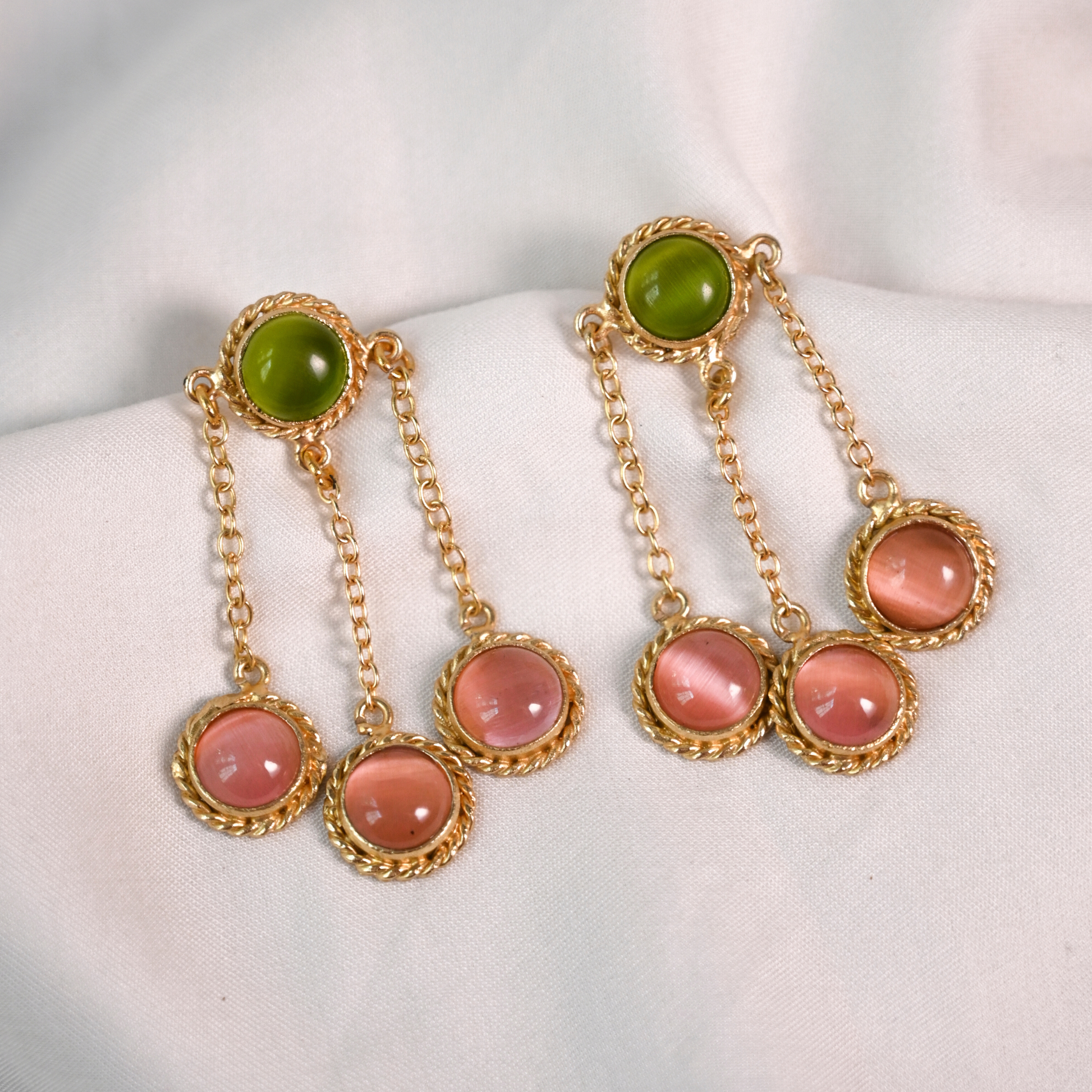 Gold-plated dangle earrings with green and translucent pink stones arranged on white fabric.