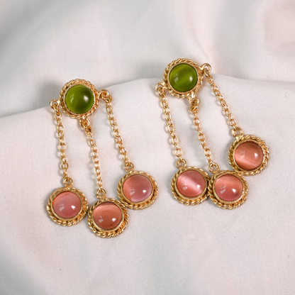 Gold-plated dangle earrings with green and translucent pink stones arranged on white fabric.